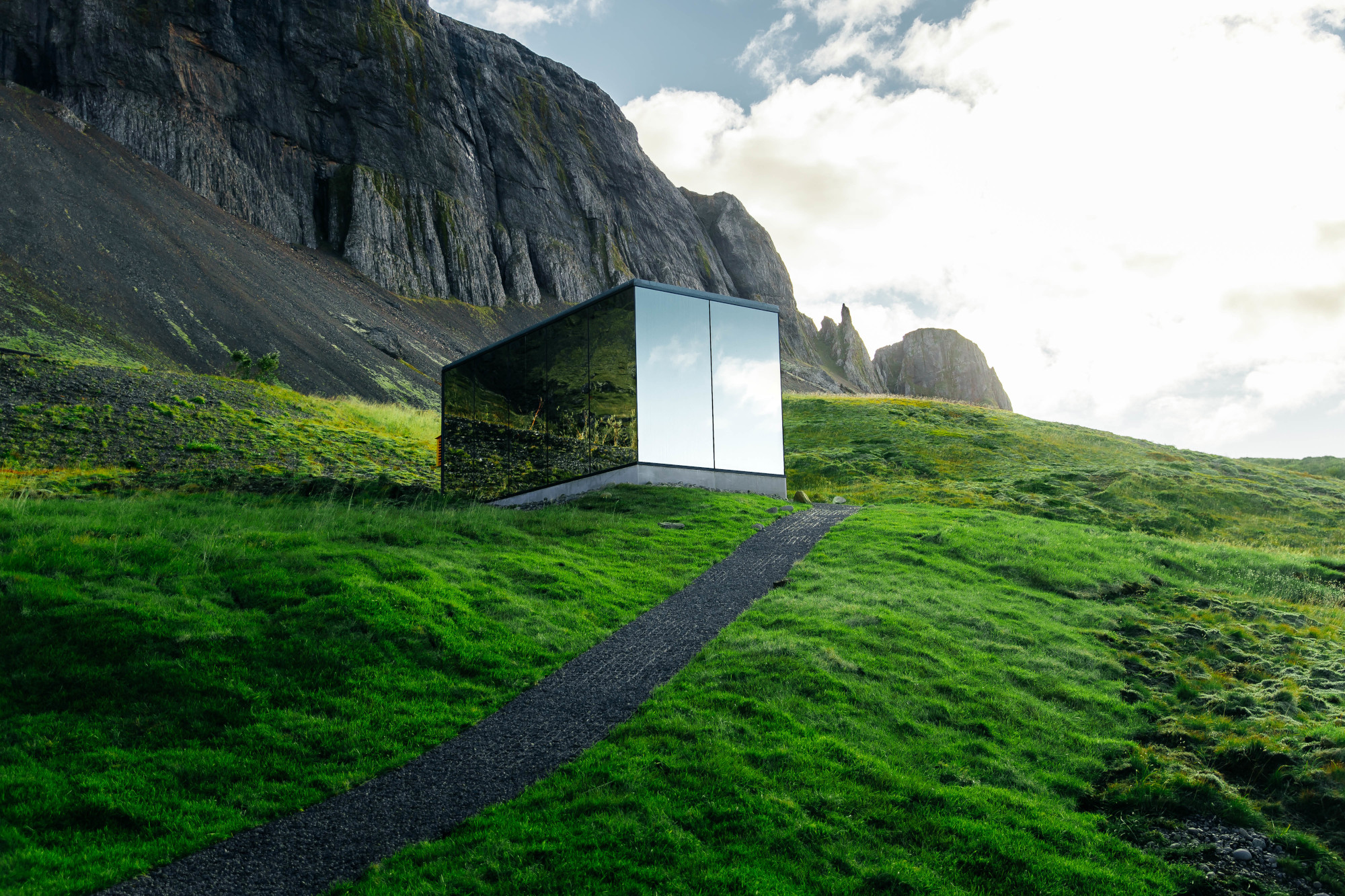 The mirrored glass cabin below a basalt cliff, reflecting the grass and sky